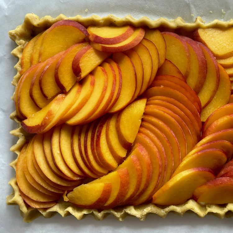 Image of Cut your peaches in half to remove the pits, then lay them flat on a cutting surface. Slice the peaches into 1/8” slices and arrange them on the prepared crust by fanning them slightly.